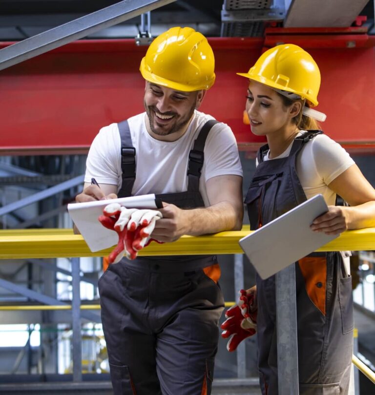 Two construction workers in yellow hard hats review a clipboard and tablet on a yellow railing in an industrial setting
