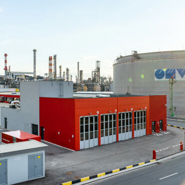 Red industrial service building with multiple garage doors at a chemical plant, large storage tanks in the background