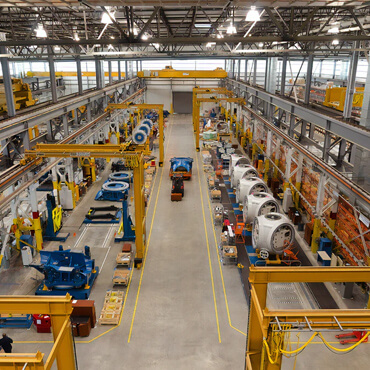 Inside a large industrial workshop with yellow overhead cranes, workstations, and large metal components on both sides of the aisle